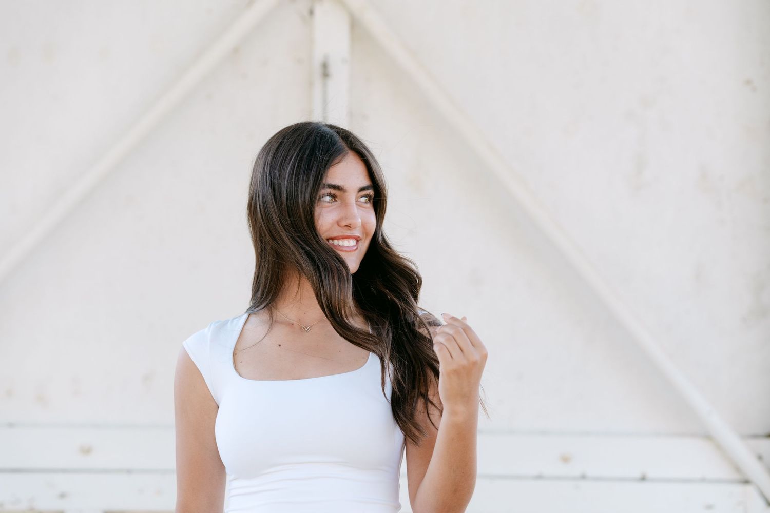 Young woman with long dark hair wearing a white tank top poses outdoors against a light-colored wall.