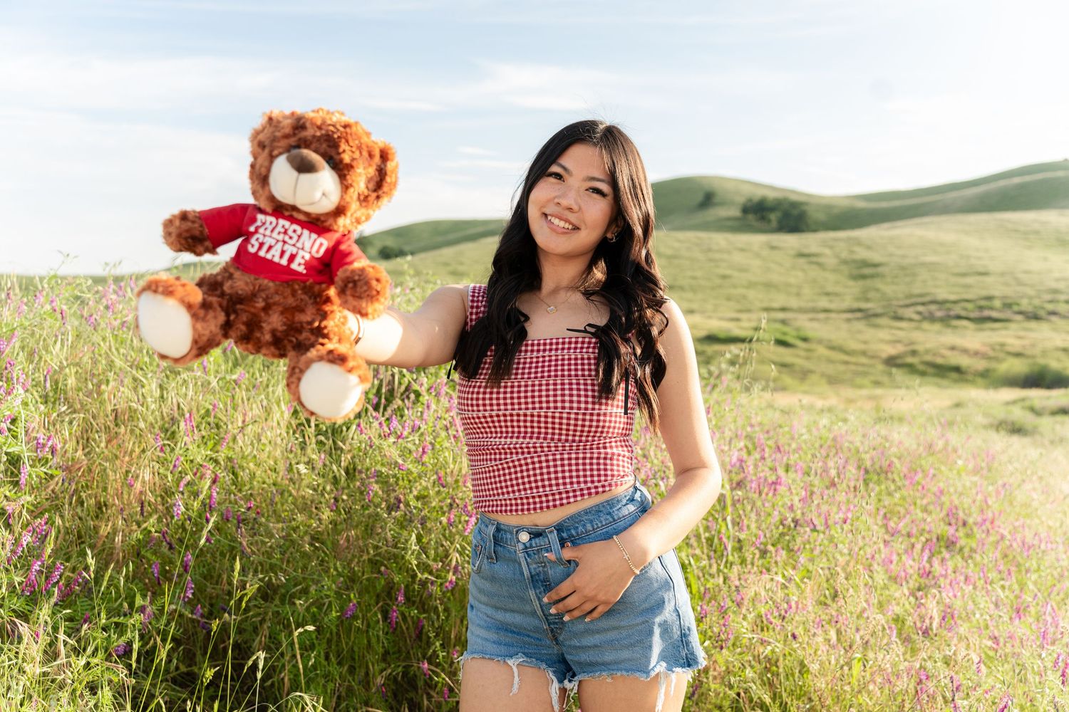 A person in a red gingham top and denim shorts holds a teddy bear while standing in a wildflower meadow.