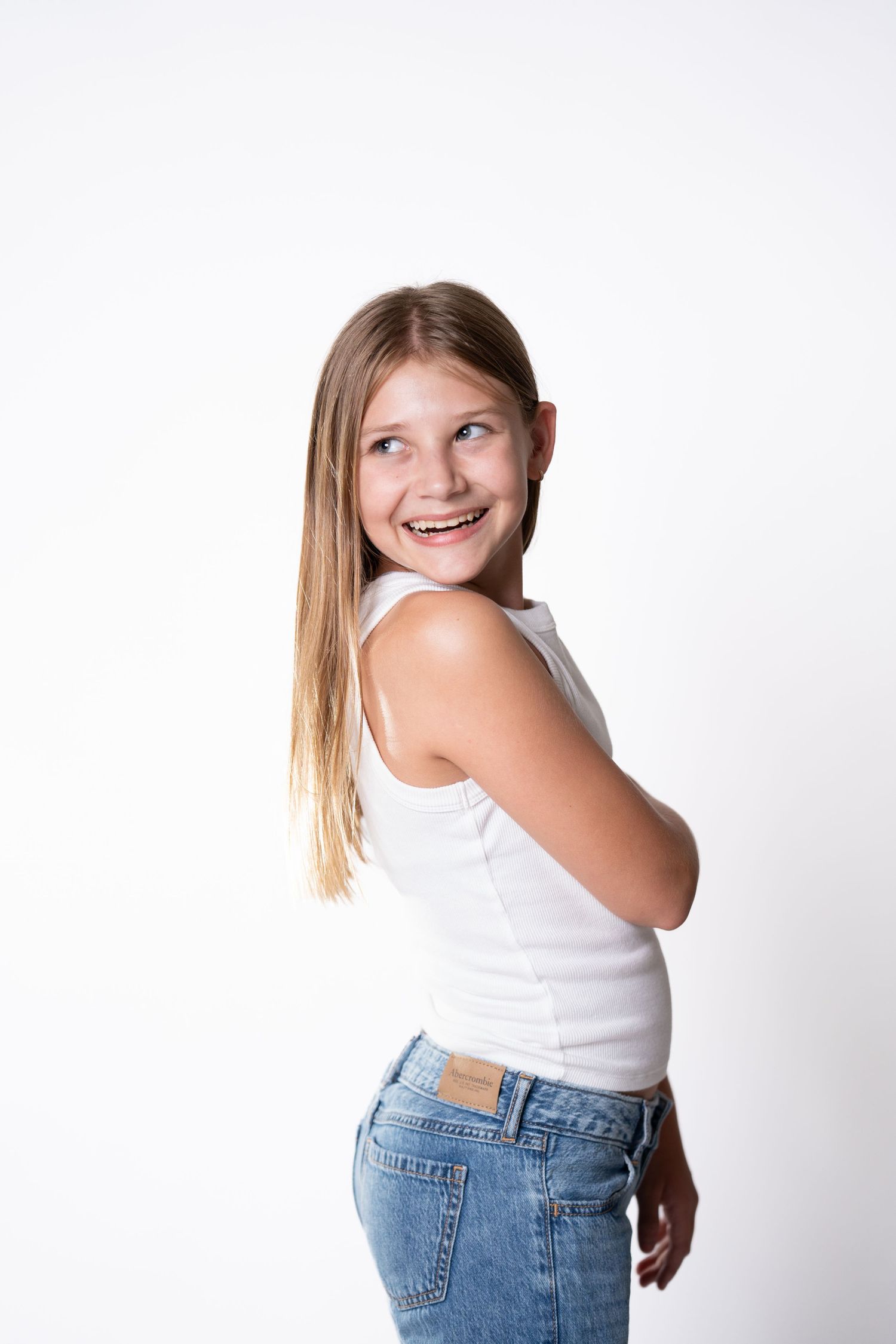 Young person in casual white tank top and jeans poses playfully against white background.