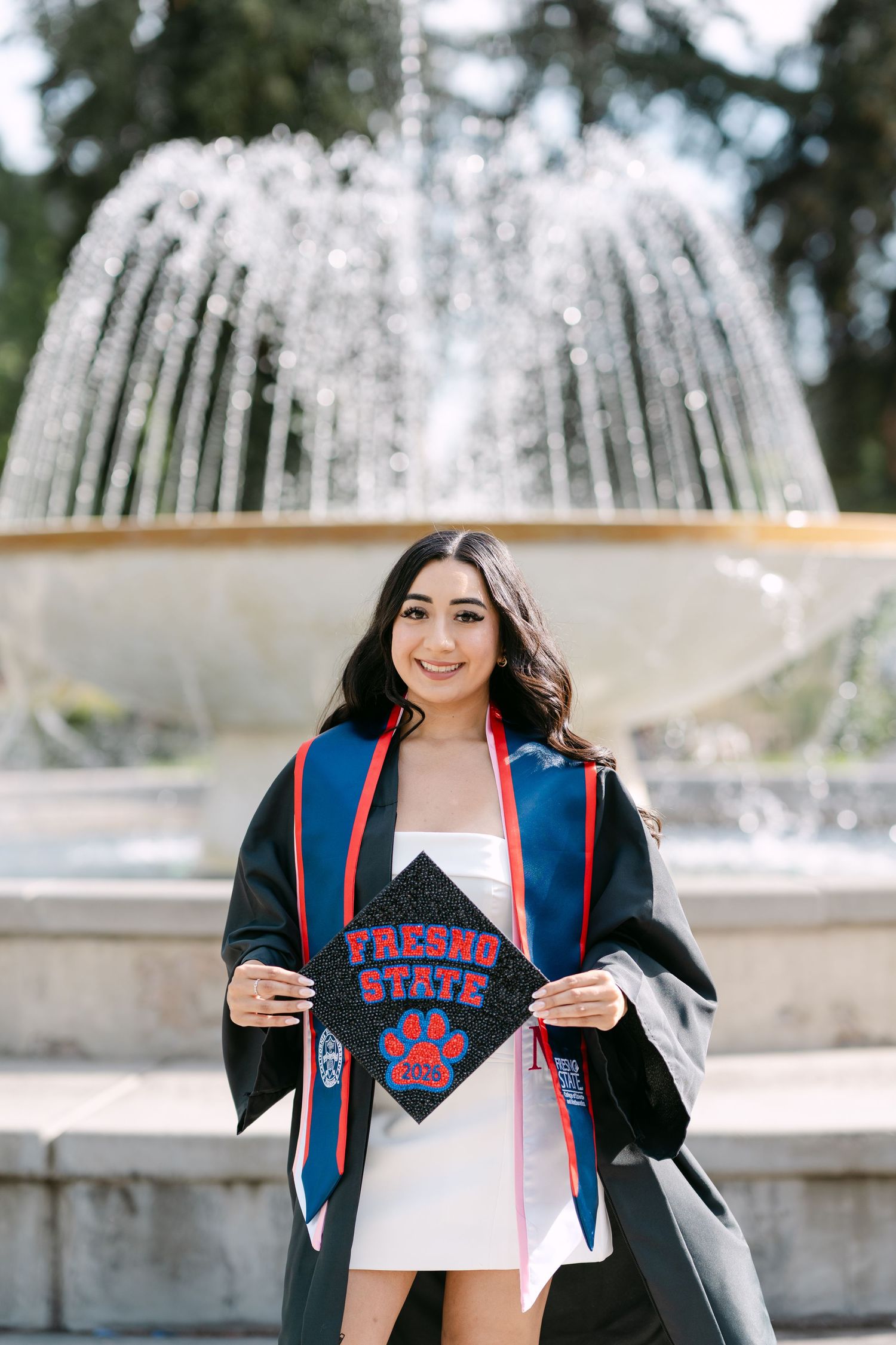Fresno State graduate smiling in cap and gown, holding decorated mortarboard, posing in front of a fountain.