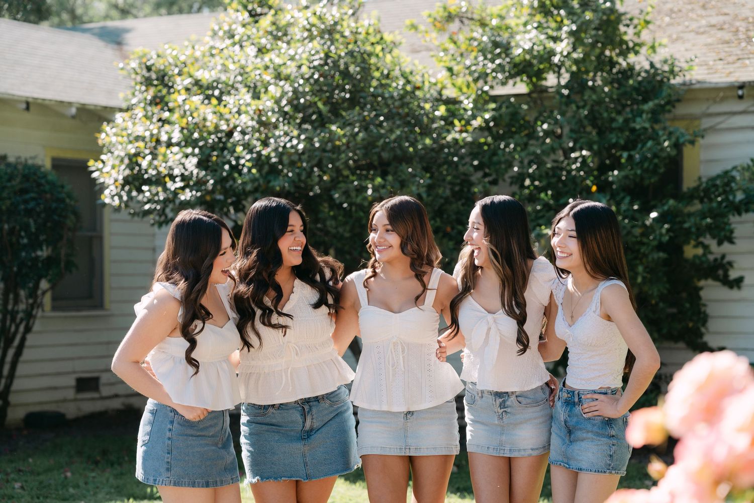 Five young women in white tops and denim skirts laughing together outdoors in a sunny garden setting.