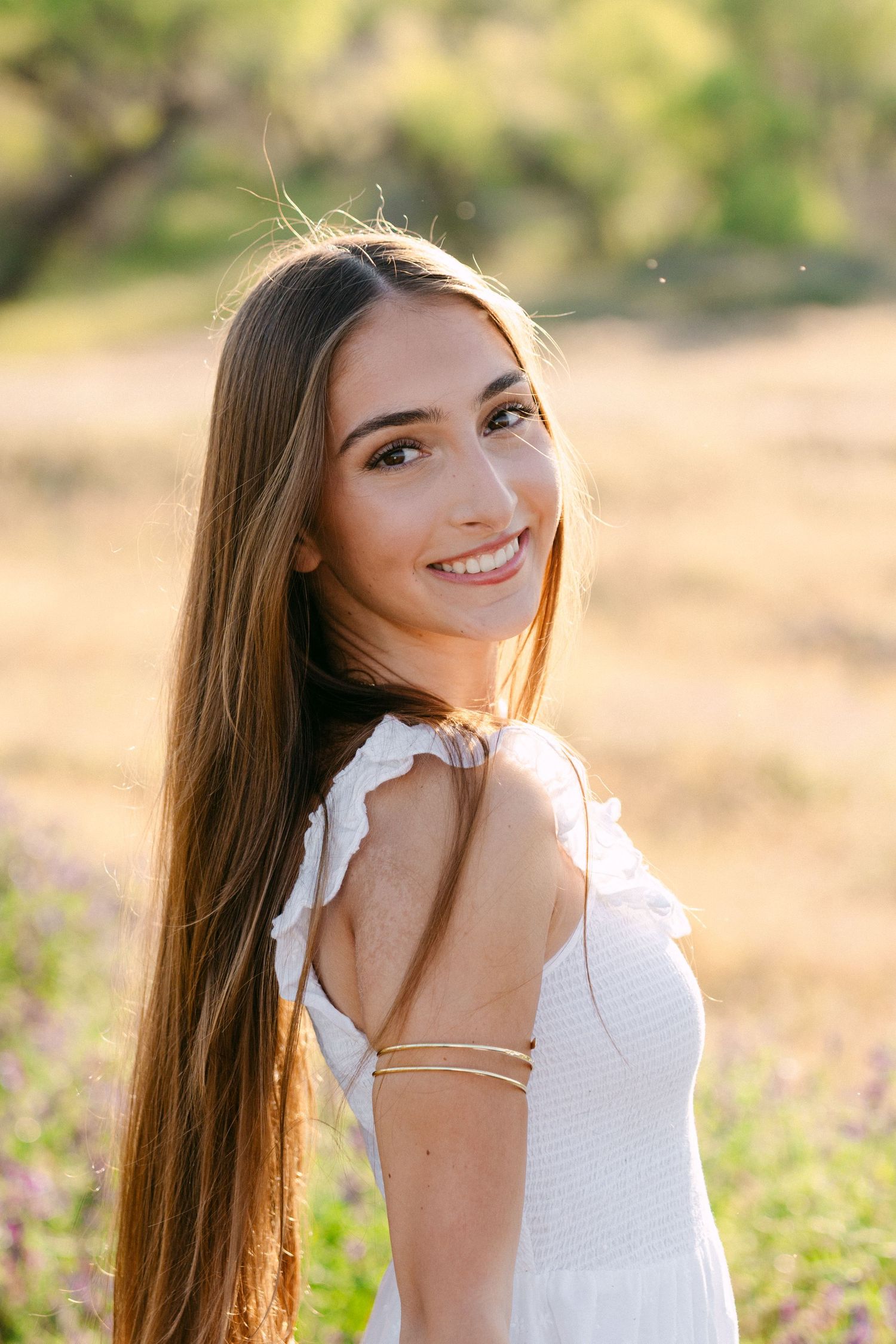 Young woman with long brown hair smiling outdoors in a white dress, golden sunlight illuminating a natural field background.