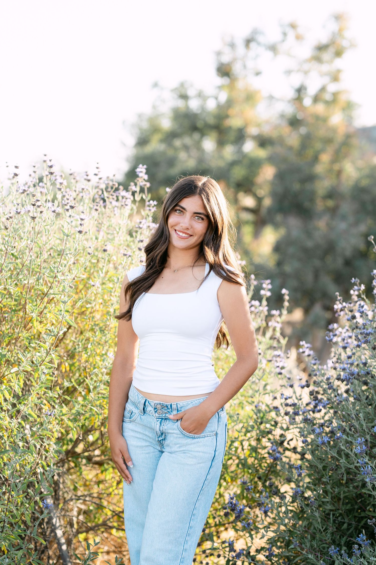 Young woman in white crop top and light jeans smiling outdoors among purple wildflowers and green trees.