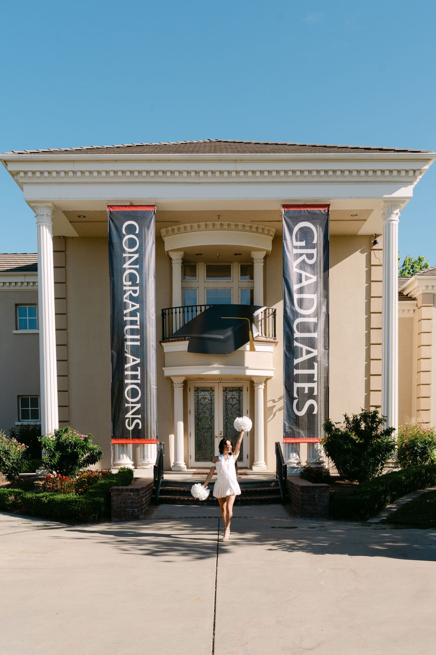 Graduate in white dress holding balloons stands before a grand building with Congratulations Graduates banners.