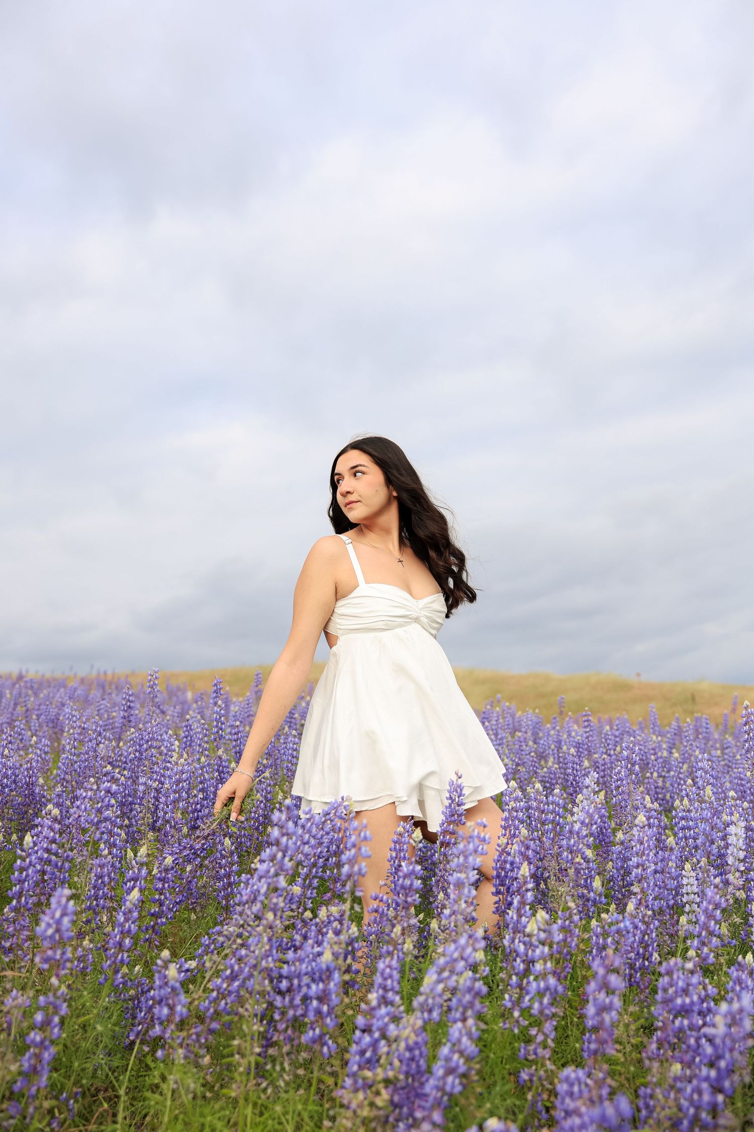 Woman in white dress walking through blooming purple lupine field under cloudy sky.