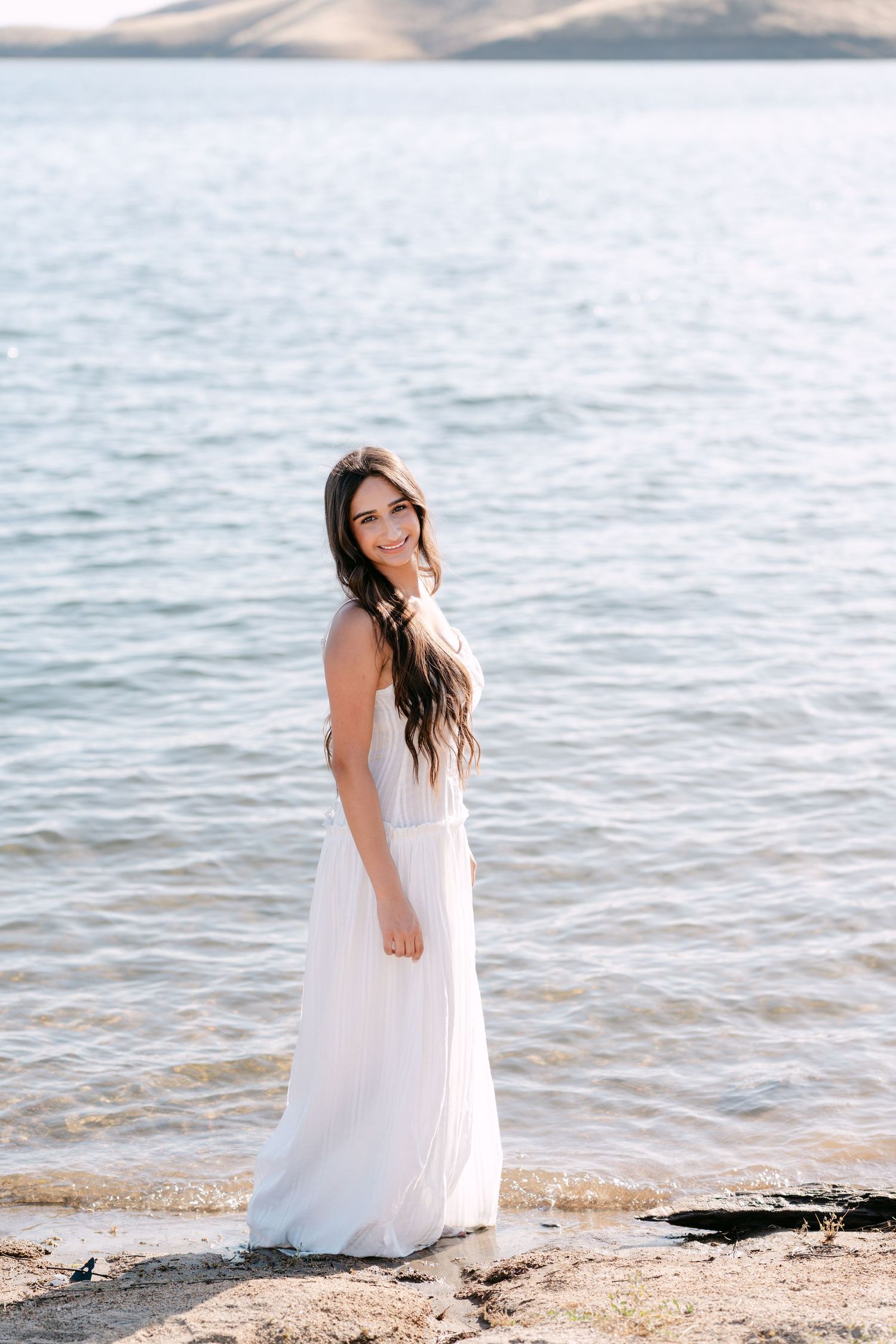 Smiling brunette woman in a flowing white dress standing on rocky shore with calm lake and mountains in background.