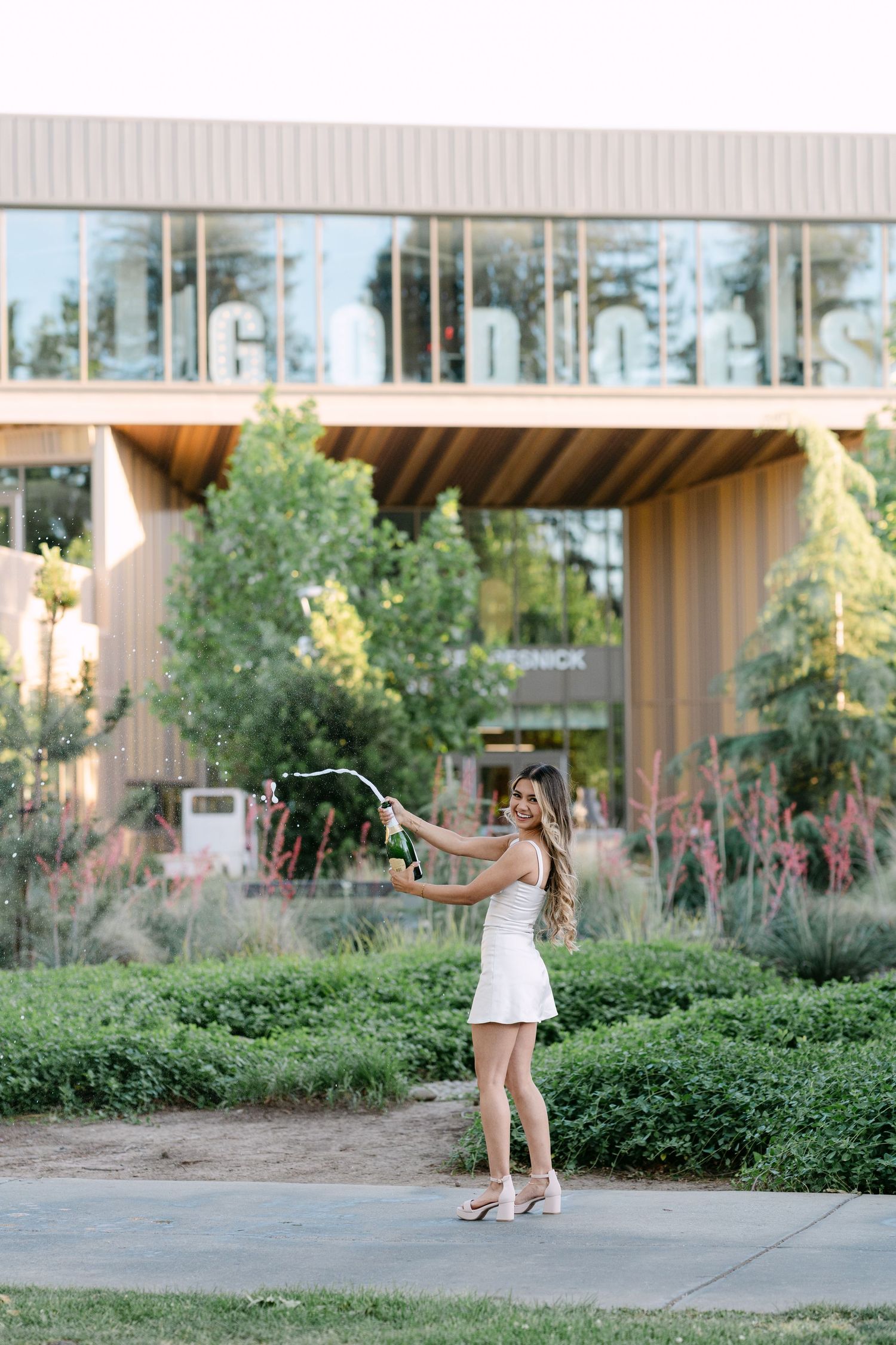 Young woman in white dress popping champagne bottle outdoors near modern building with lush greenery.