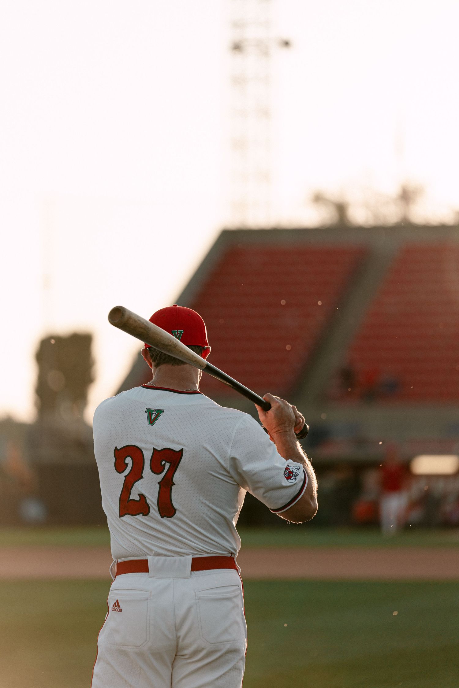 Baseball player in white uniform swings bat during sunset practice at stadium with red bleachers visible in background.