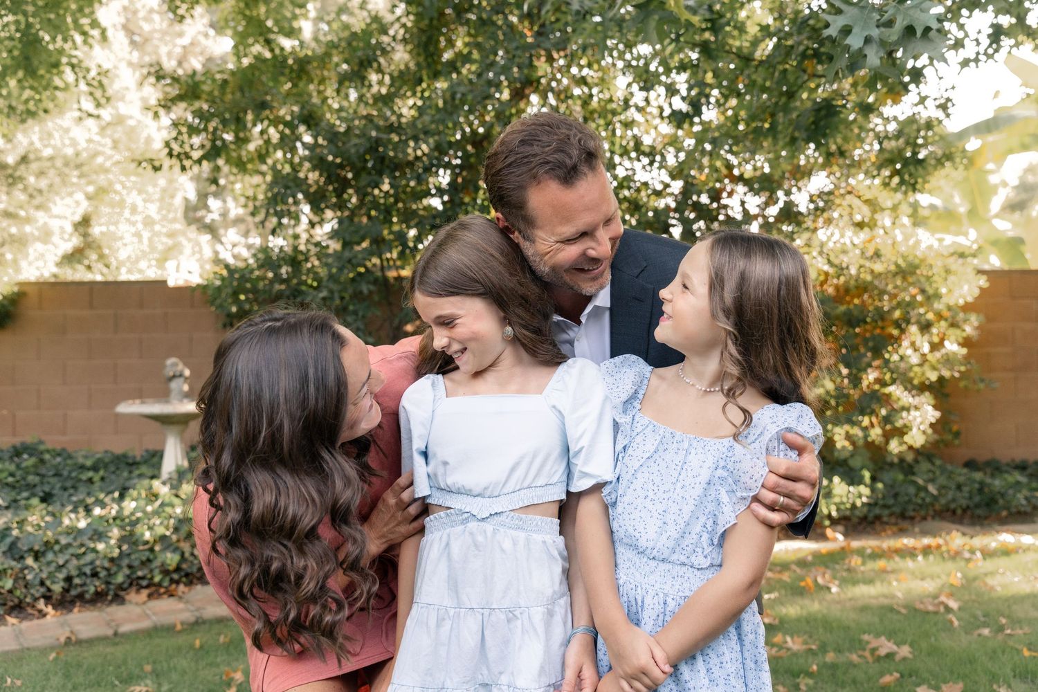 A family of four shares a joyful moment together in a sunlit garden with greenery in the background.