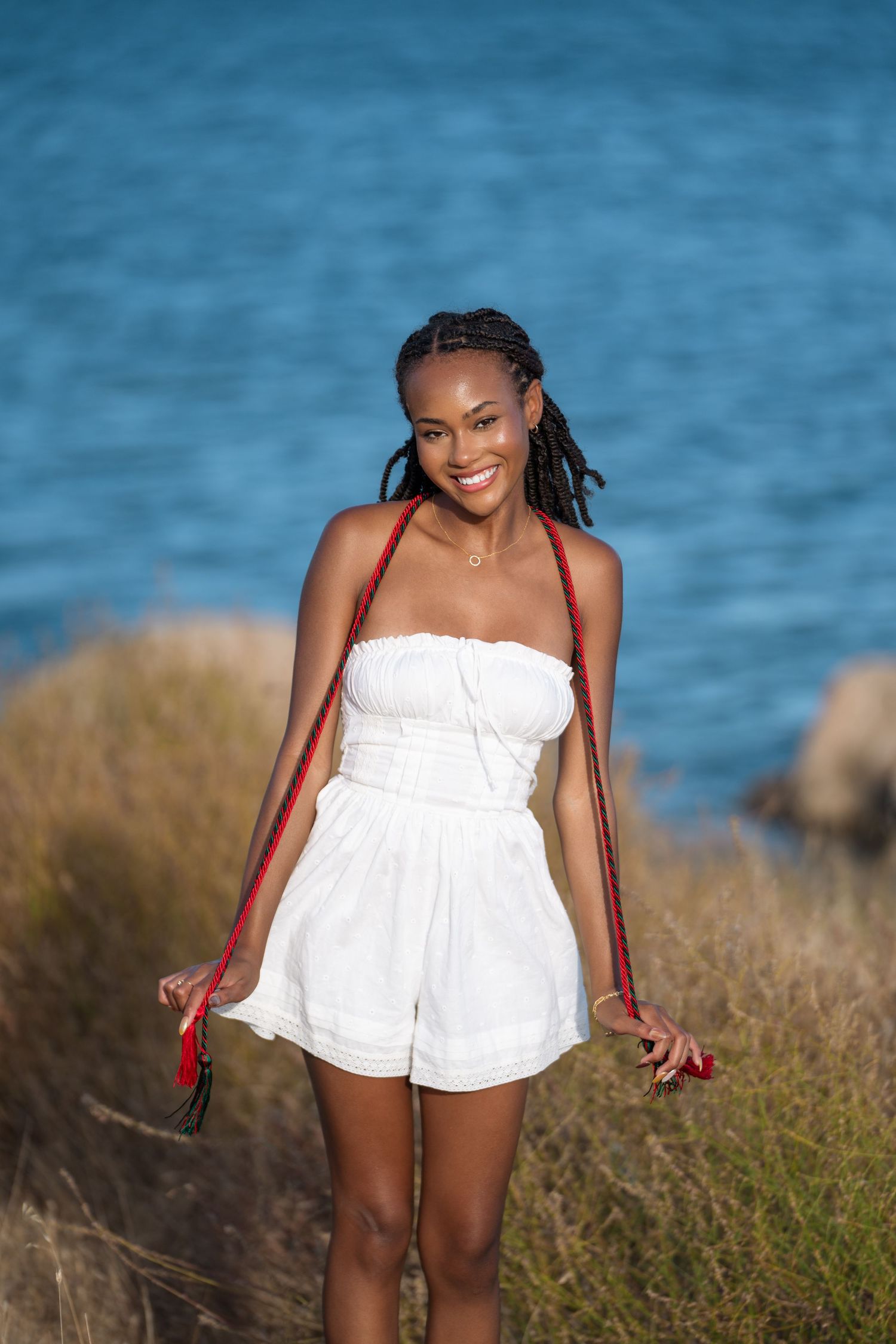 Smiling young Black woman in white strapless romper with red braids stands near blue water on a sunny day.