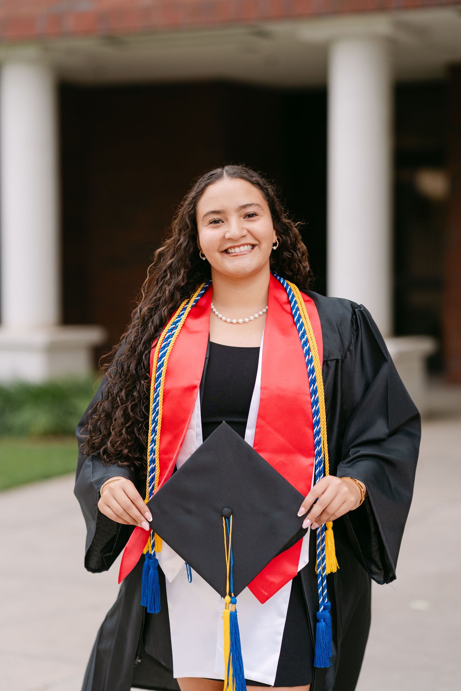 Smiling female graduate in black gown holding cap with colorful honor stoles outdoors near columned building.