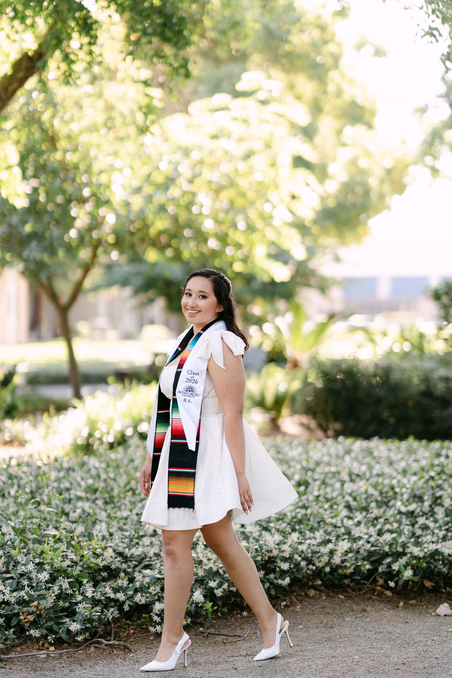 Smiling graduate in white dress and colorful sash walks outdoors among green trees and white flowers.