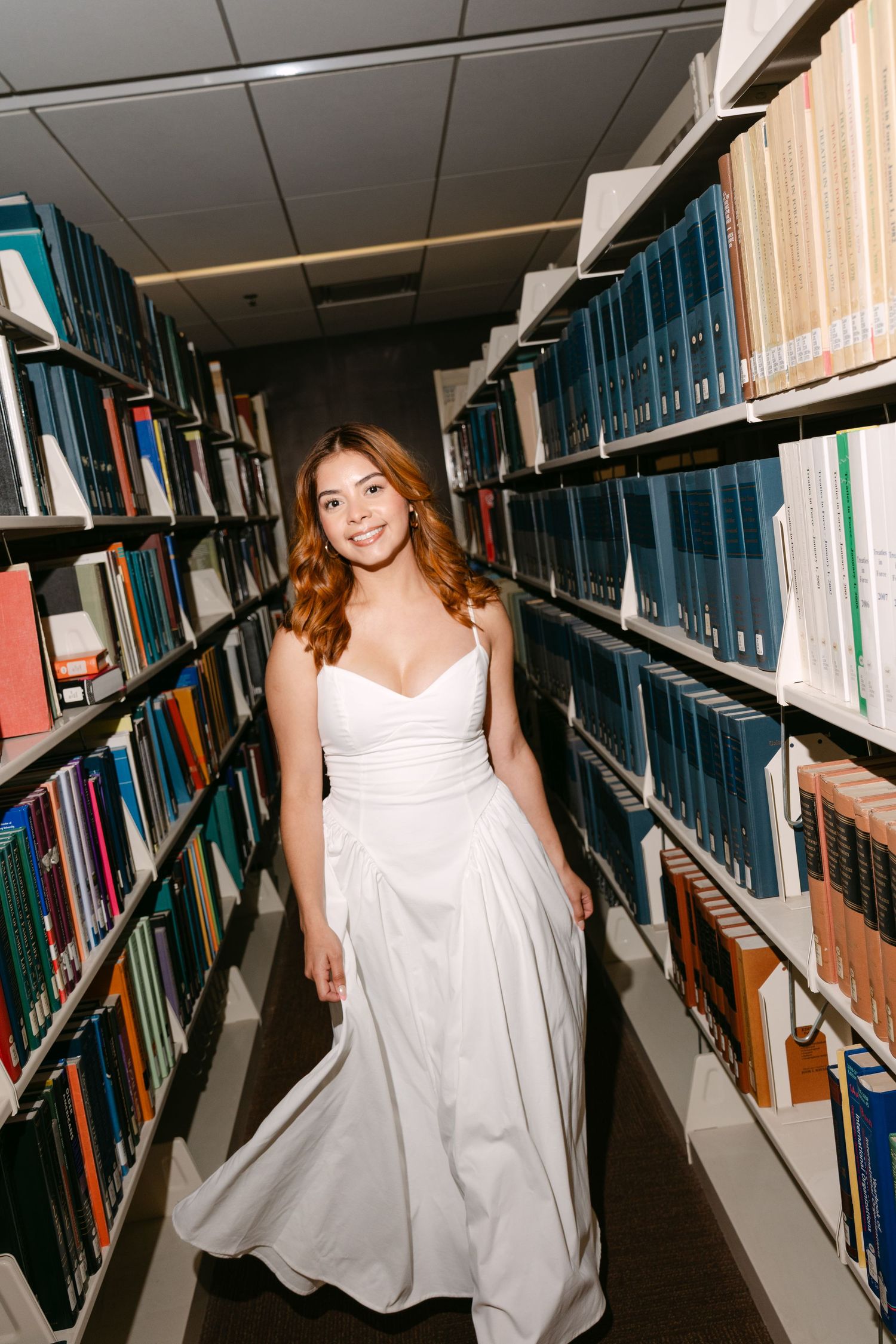 A smiling bride in a white gown walks between library bookshelves in an elegant pre-wedding photoshoot.