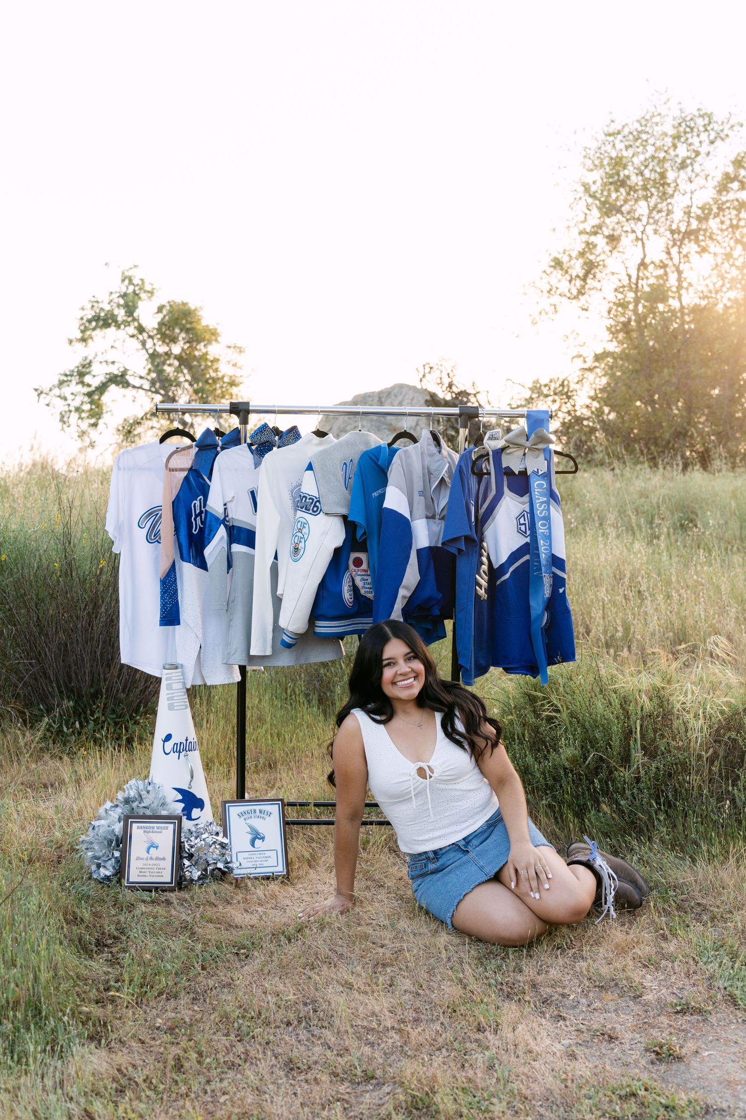 Smiling young woman sitting outdoors beside a clothing rack displaying blue and white cheerleading uniforms at golden hour.