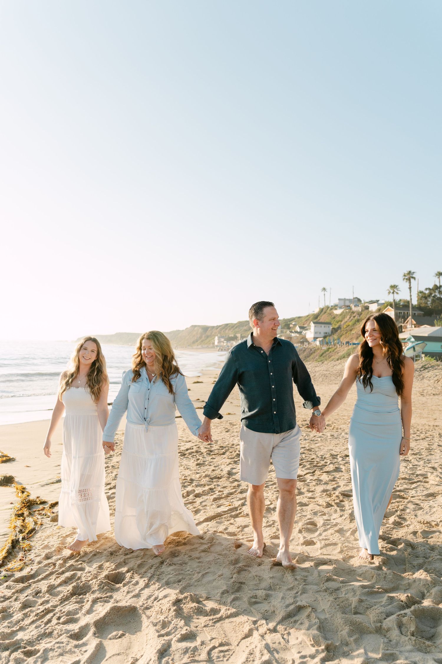 Family of four holding hands walking on a sunny beach, wearing light blue and white outfits with ocean views.