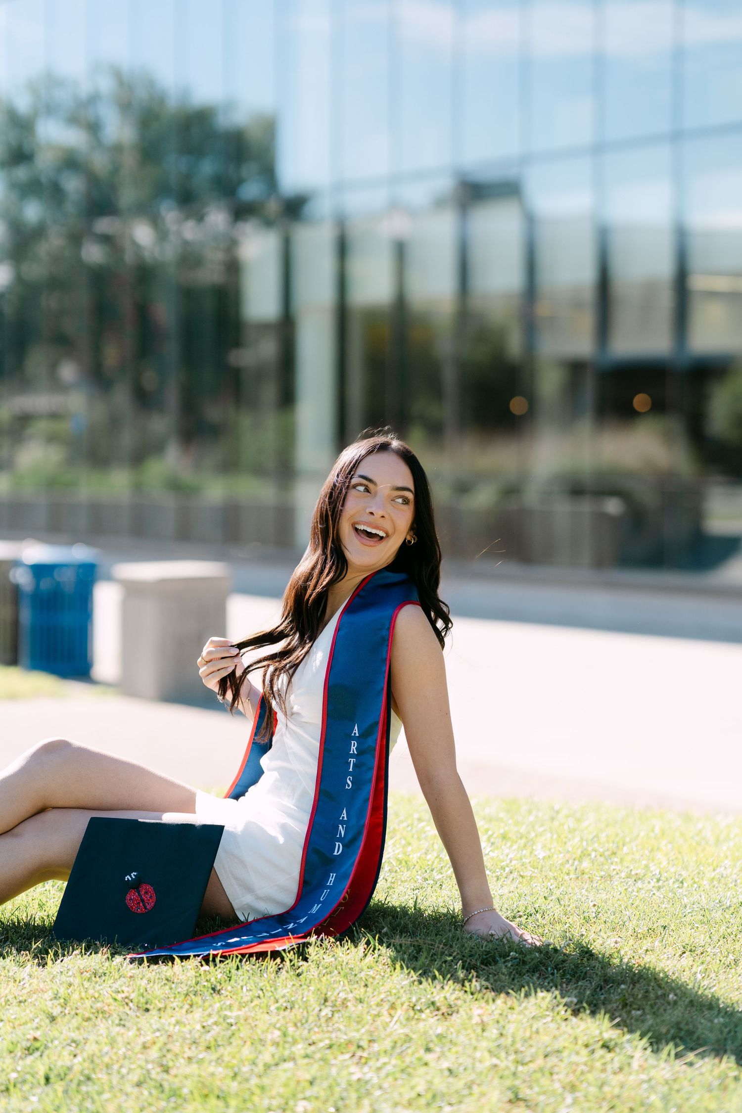 Smiling female graduate sitting on grass wearing white dress and navy blue graduation stole with mortarboard cap beside her.