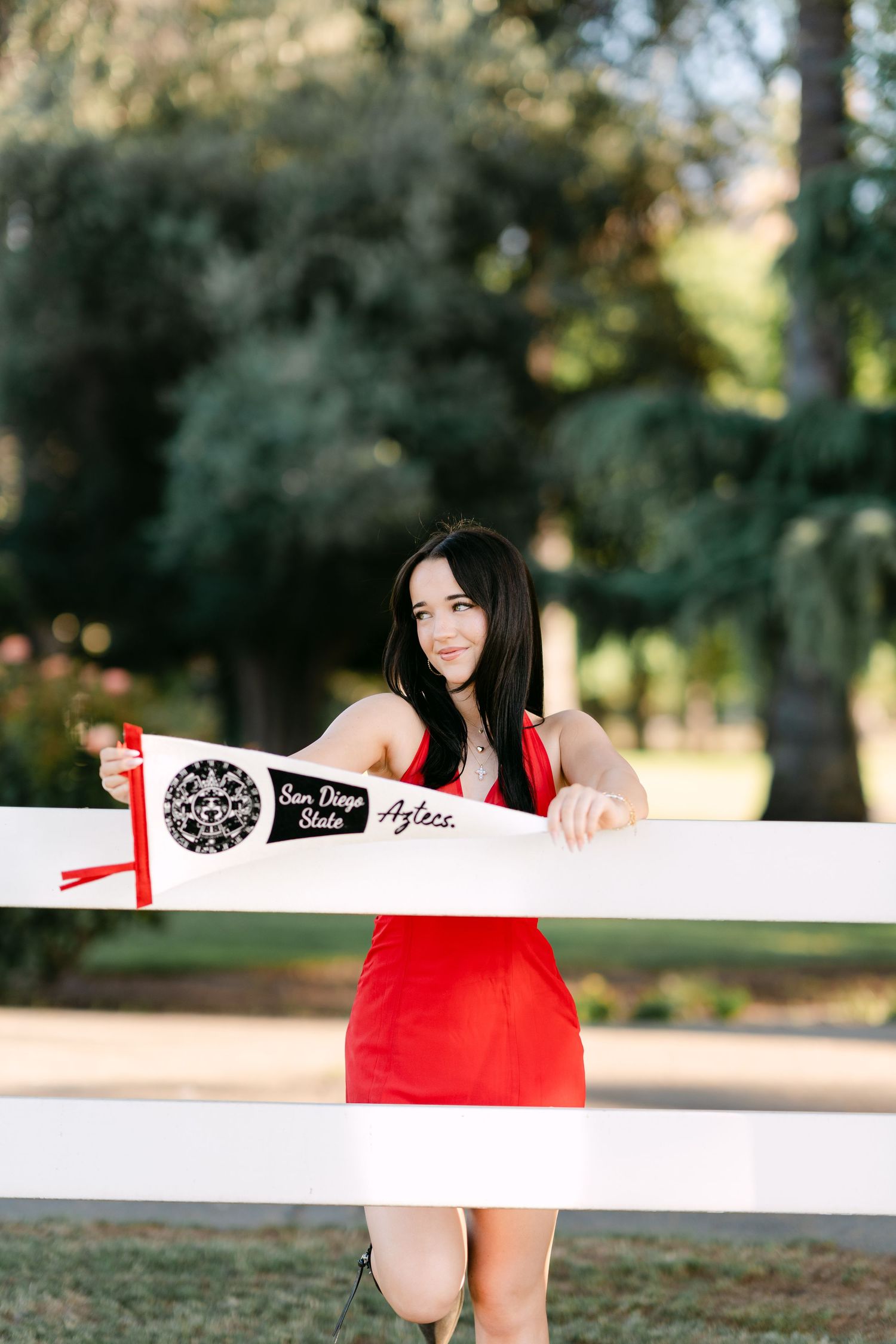 Young woman in red dress holding San Diego State Aztecs pennant flag while leaning on white fence outdoors.