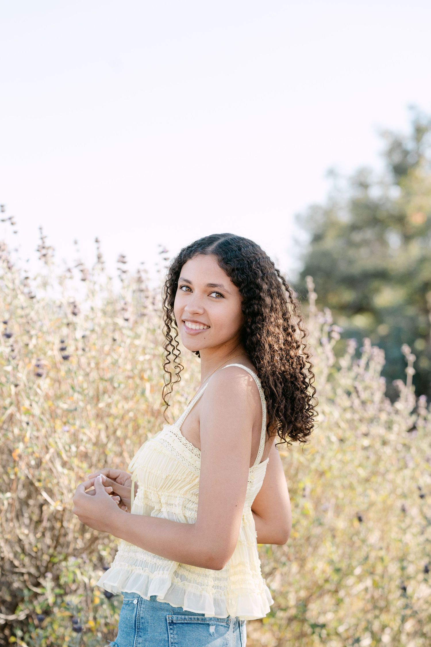 Young woman with curly hair smiles outdoors in a yellow top and jeans, surrounded by wildflowers in golden light.