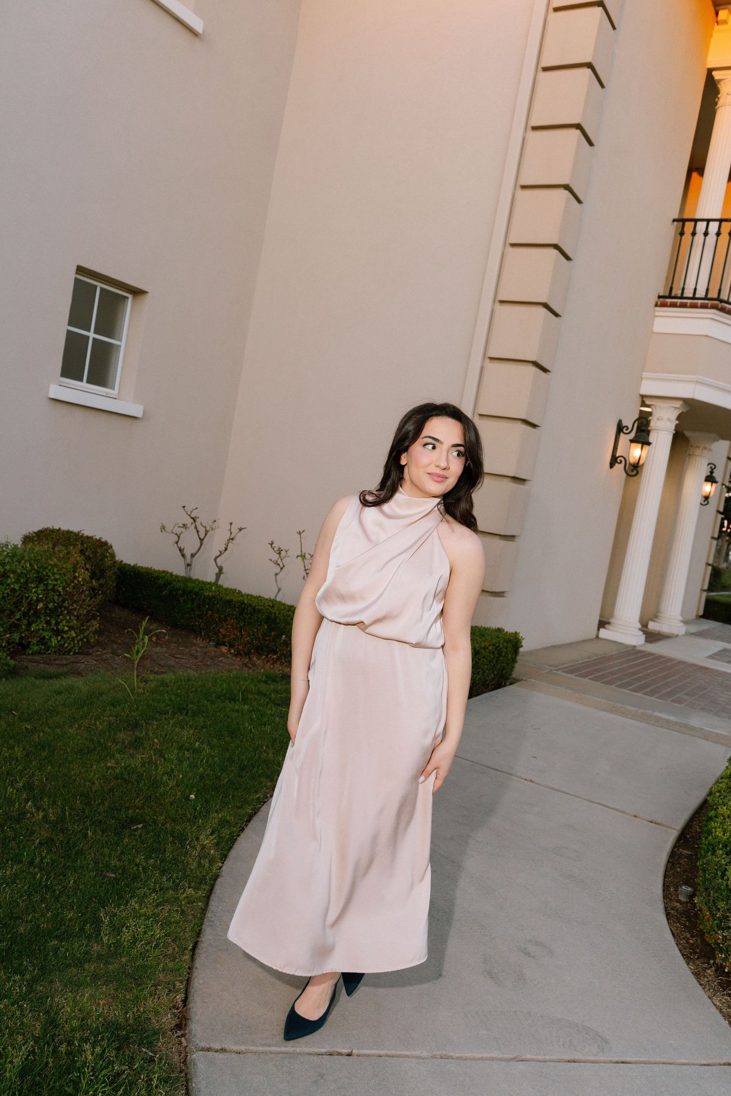 A person in a light pink sleeveless dress poses in front of a cream-colored building exterior.