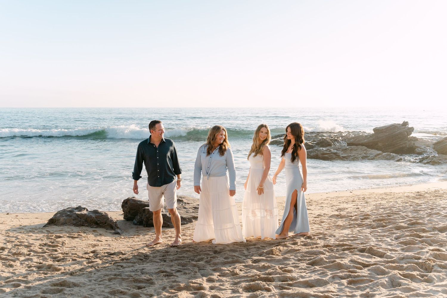 Family of four walking on sandy beach at sunset, wearing white and navy outfits with ocean waves in background.