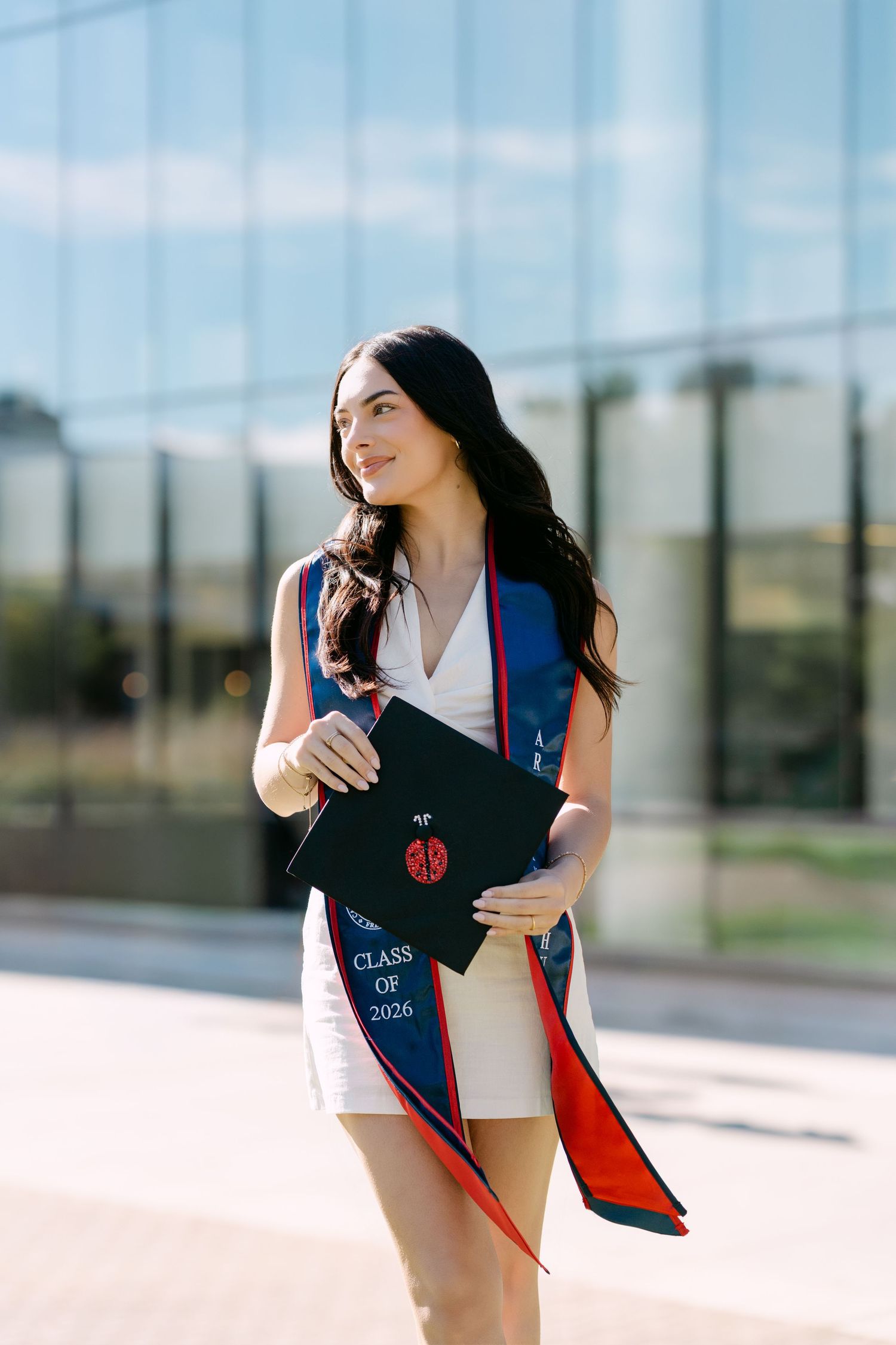 Young woman in white dress holding Class of 2026 graduation cap with blue and red stole outdoors.