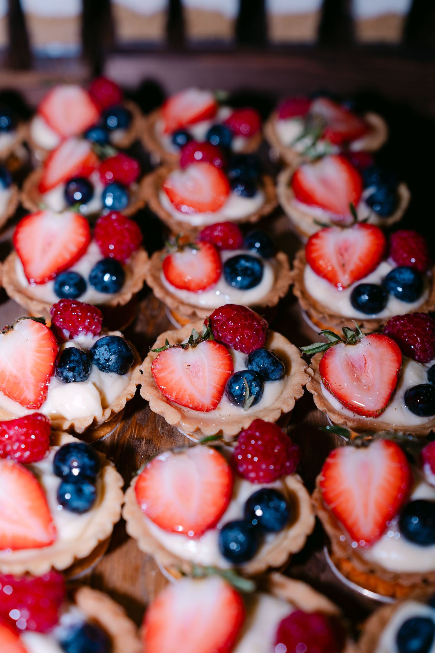Delicate fruit tarts topped with fresh strawberries and blueberries arranged in rows on a dark surface.