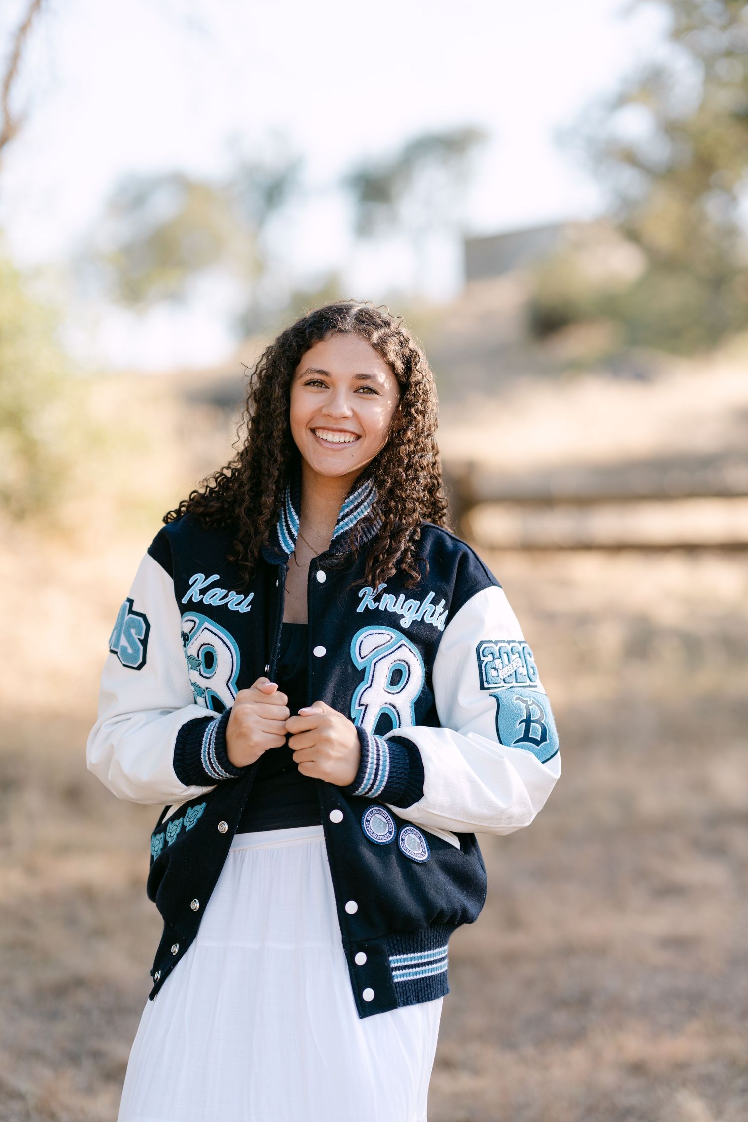 Smiling young woman wearing a varsity letterman jacket outdoors, with curly hair and a white skirt.