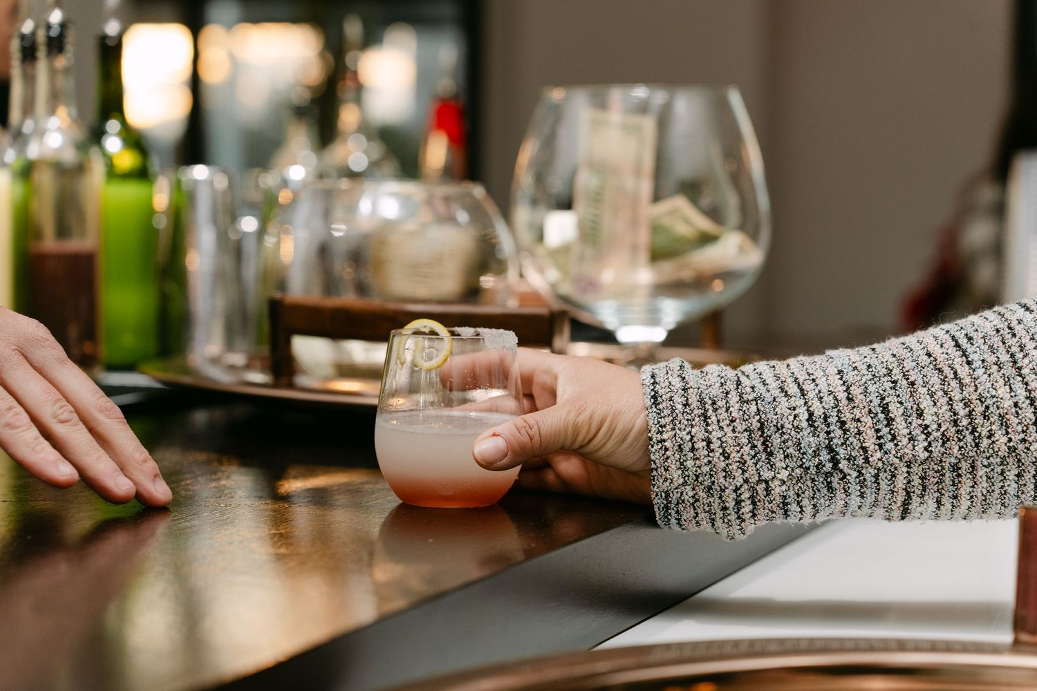 Hands reaching for cocktail glasses across a wooden bar counter.