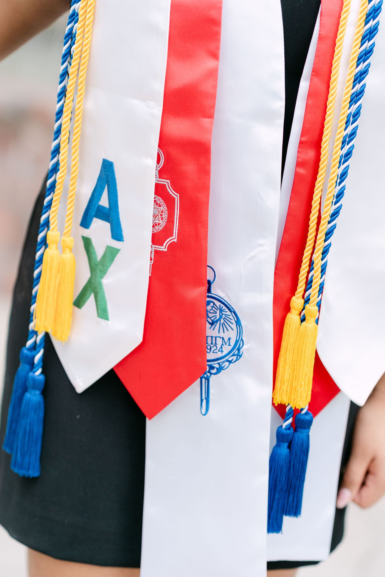 Graduation stoles and honor cords in red, white, blue, and yellow with Greek letters and tassels worn by a graduate.