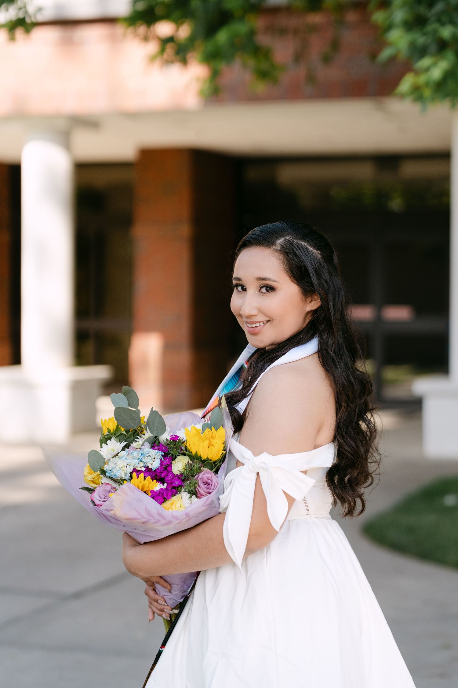 Smiling young woman in white off-shoulder dress holding colorful flower bouquet outdoors near brick building.
