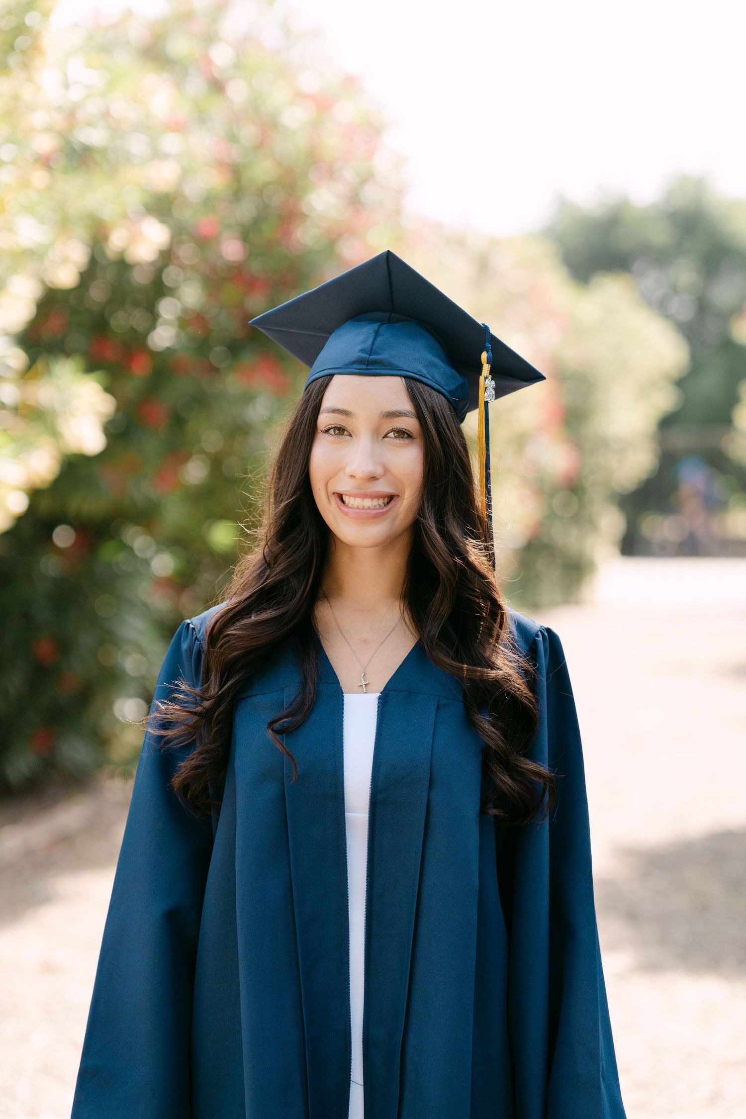 Smiling young woman in navy blue graduation cap and gown posing outdoors with colorful foliage background.