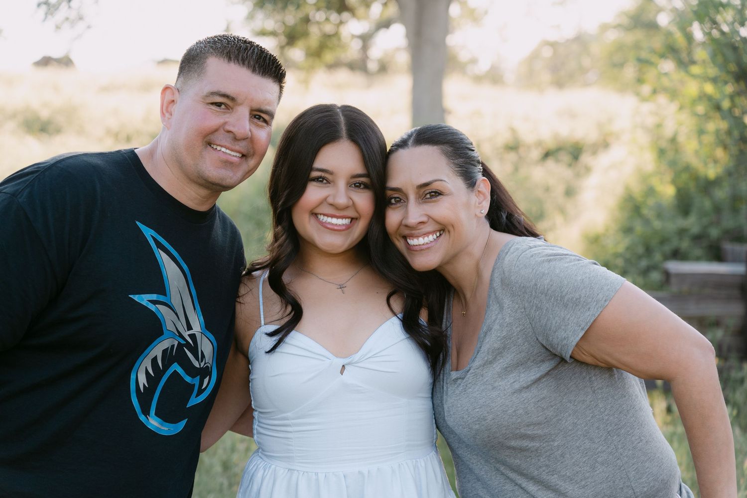 A joyful family of three smiling outdoors, with a young woman in white dress flanked by her parents.