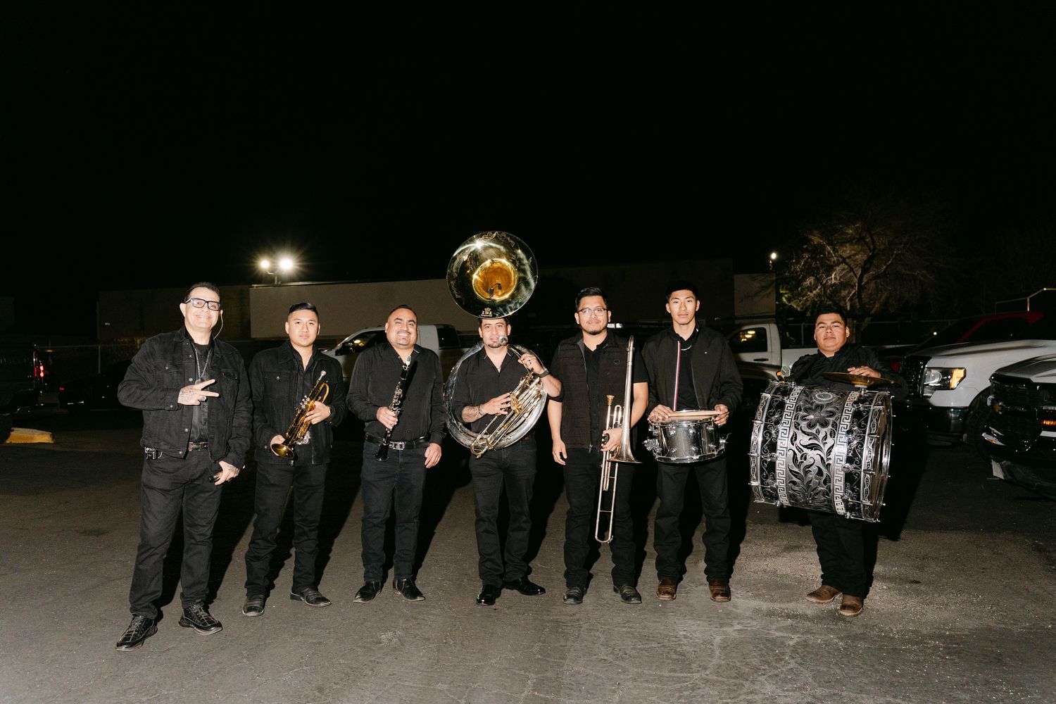 A group of musicians in black formal attire pose together with brass and percussion instruments at night.