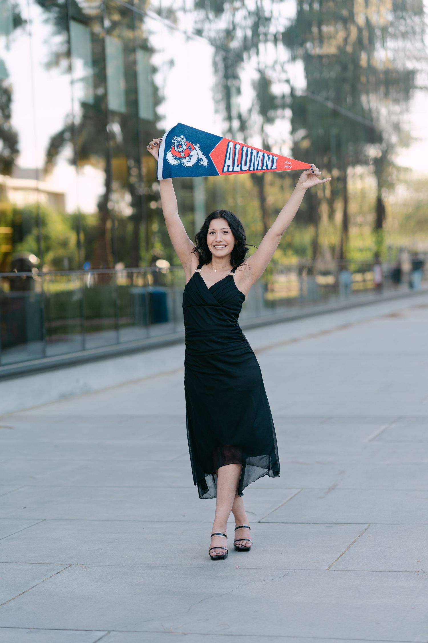 A smiling woman in a black dress holds a Fresno State Alumni pennant above her head outdoors.