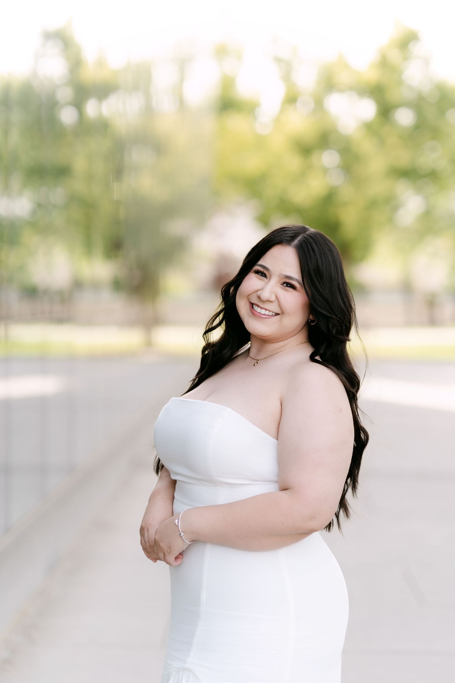 Smiling bride in strapless white gown poses outdoors with lush green trees in soft natural light.