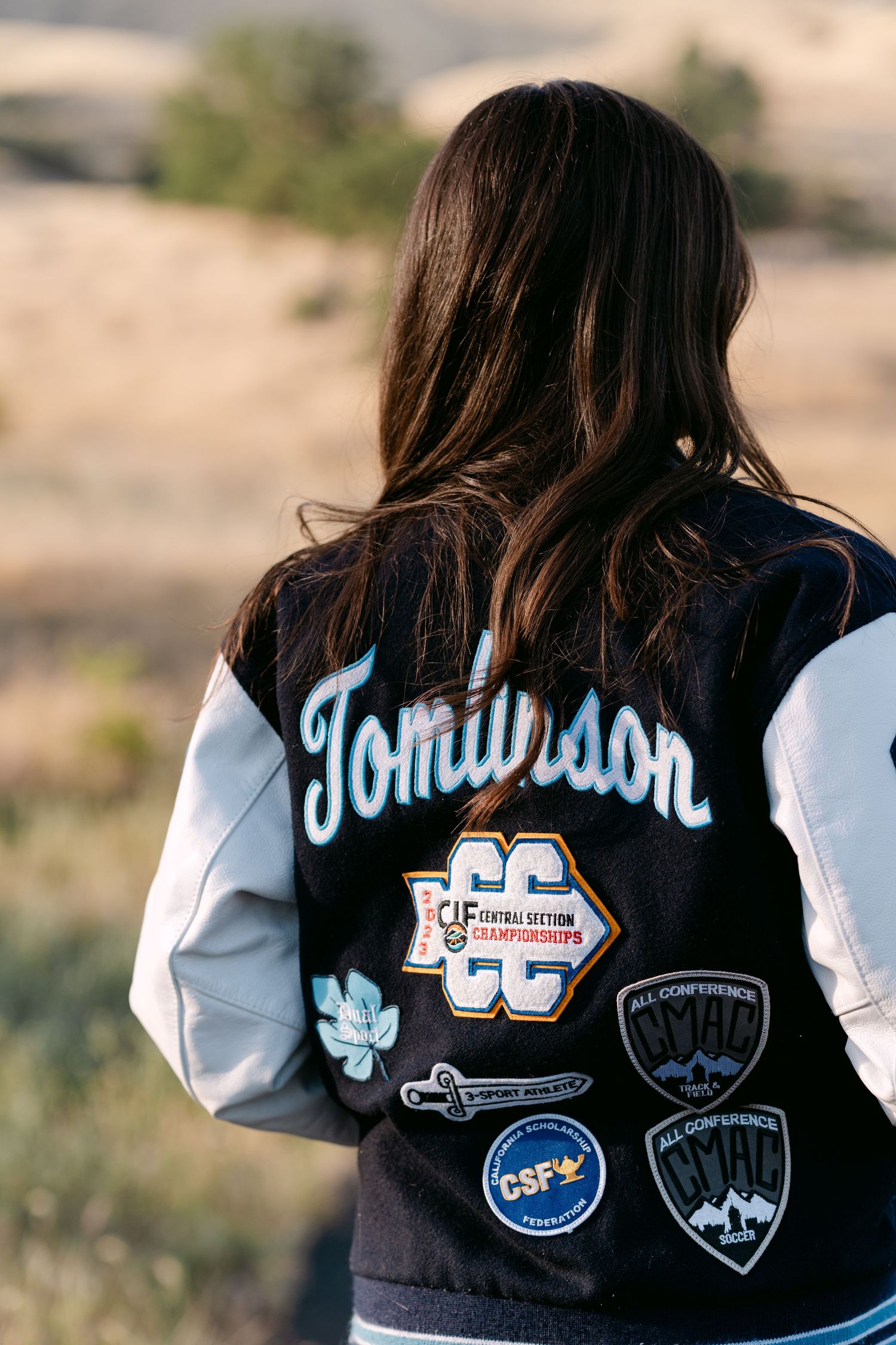 Woman wearing a varsity letterman jacket with Tomlinson name and CC Championships patches outdoors.