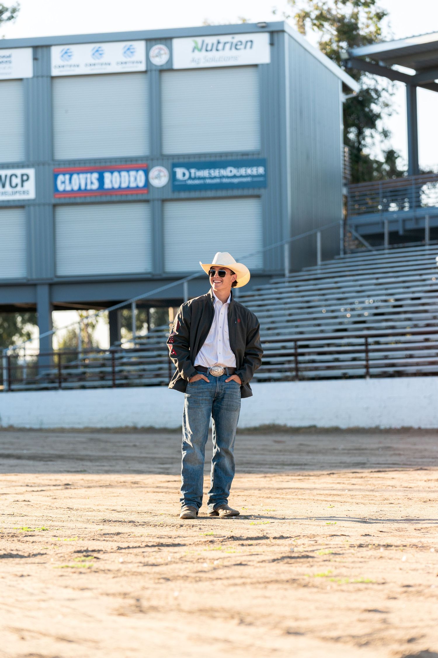 A photographer in a cowboy hat and jacket captures shots at a rodeo arena during golden hour lighting.