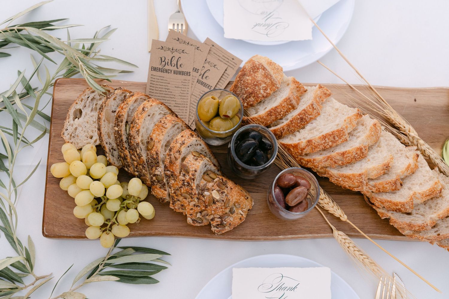 Rustic wedding reception table with bread, grapes, and olive branches on white linens.