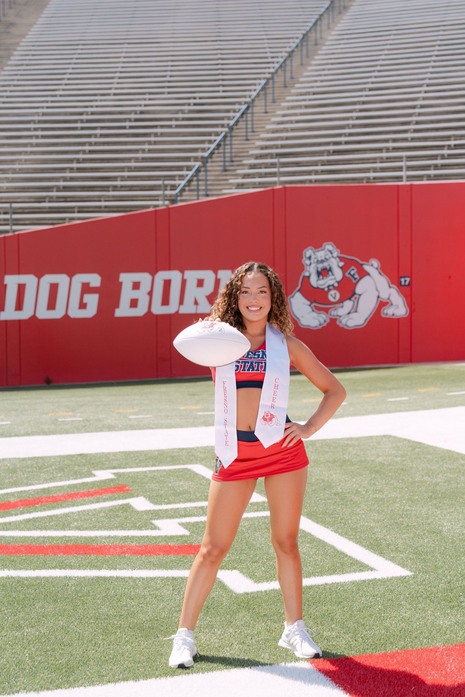 Fresno State cheerleader poses with football on field at Bulldog Stadium wearing red skirt and blue crop top.
