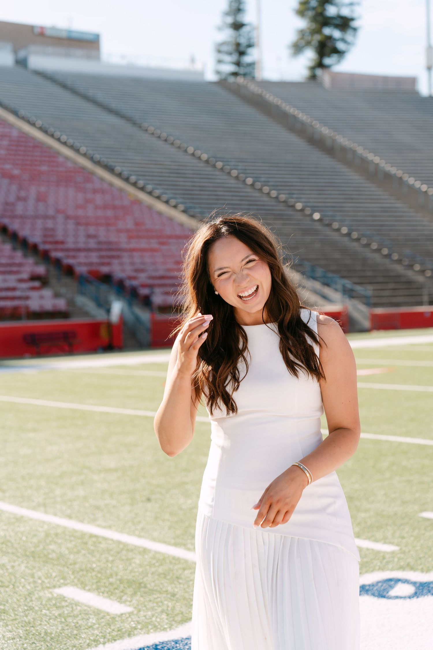 A smiling young woman in a white dress poses on a football field with stadium bleachers in the background.