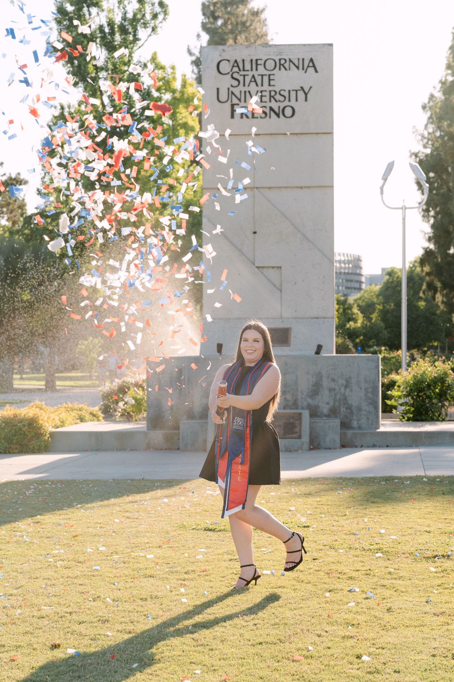 Graduate celebrating in confetti at California State University Fresno sign outdoors.