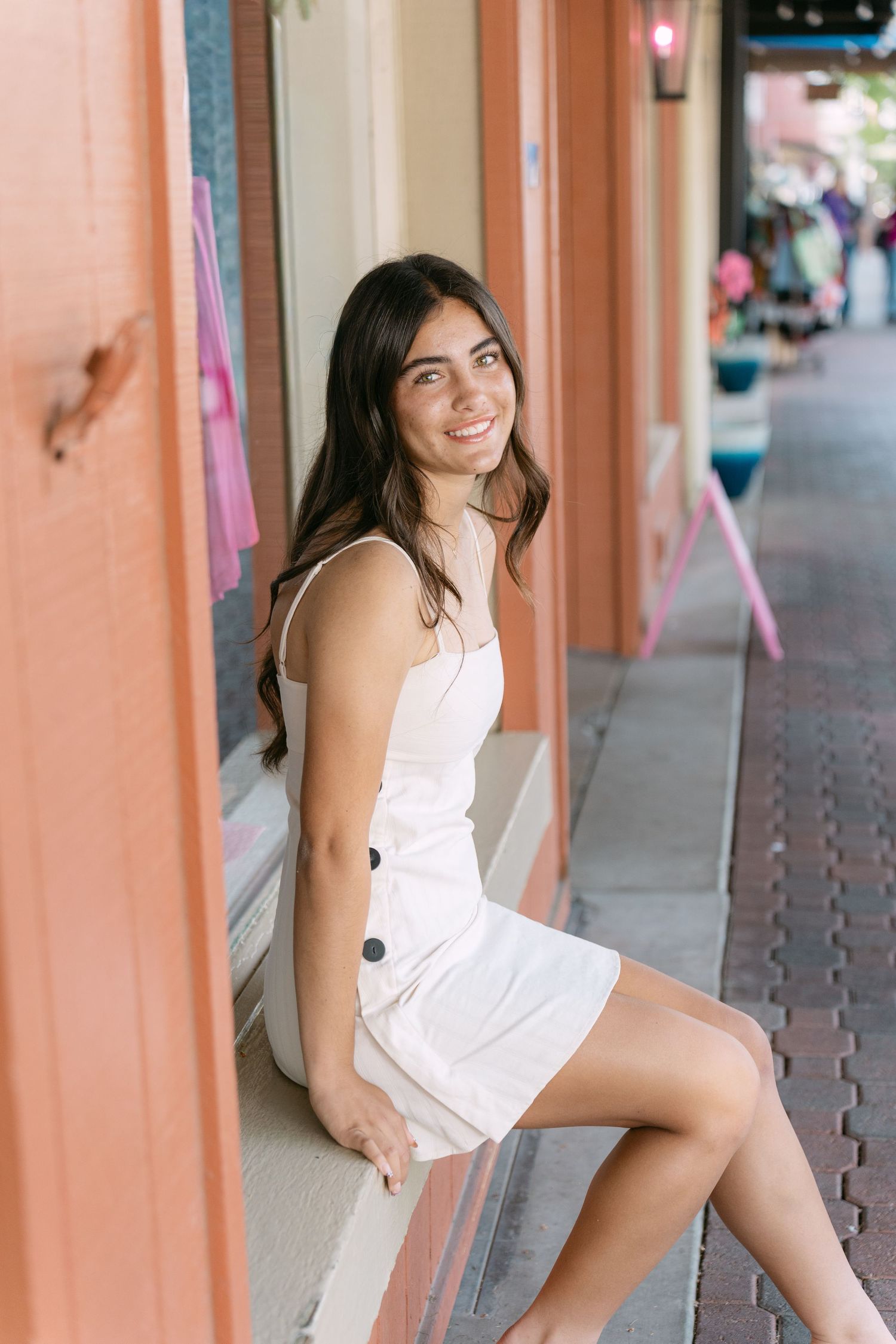 A woman in a white summer dress poses against a pink wall in an outdoor shopping area.