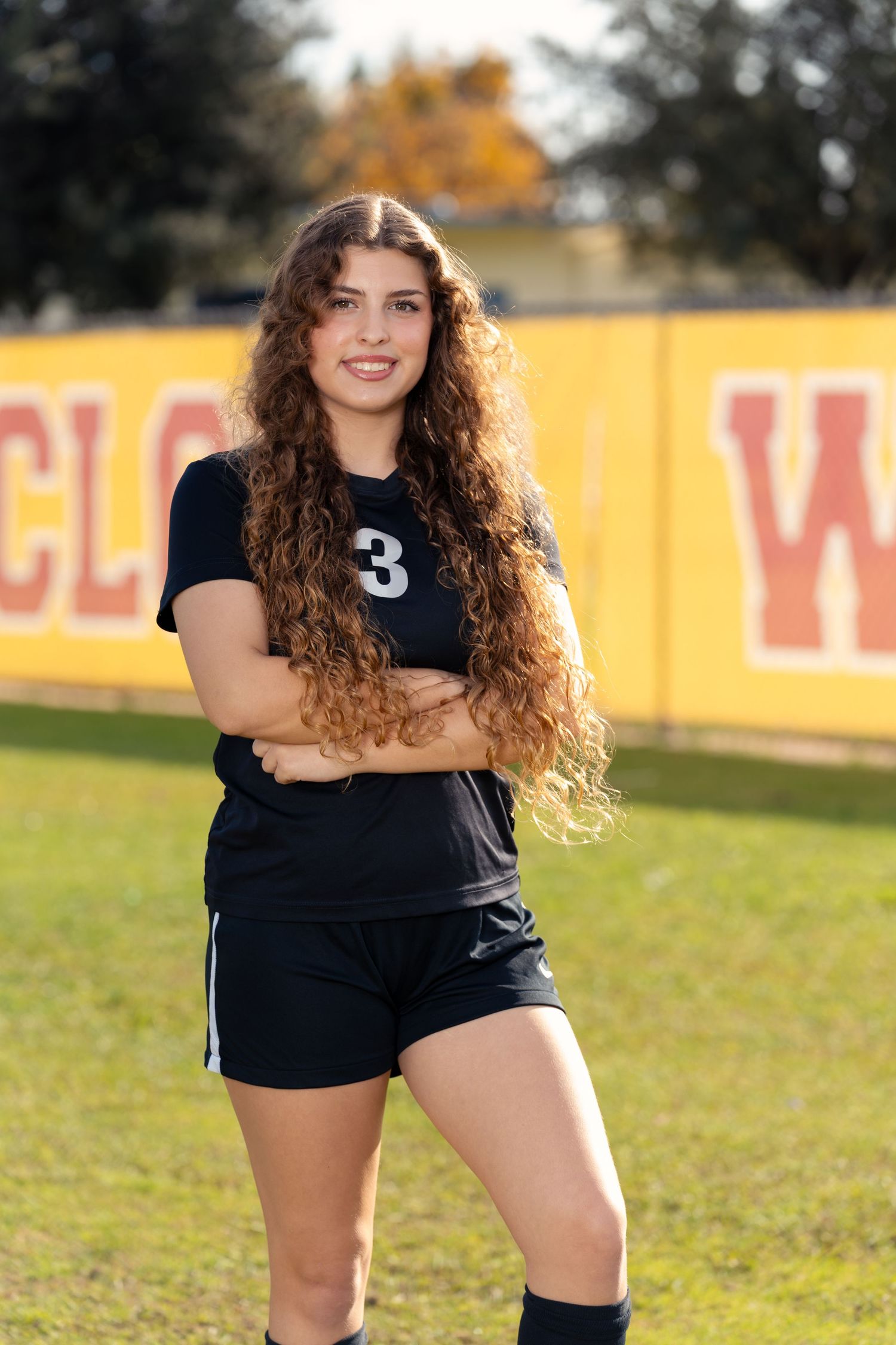 Athlete in black uniform crouches on sports field with autumn trees in background during outdoor practice session.