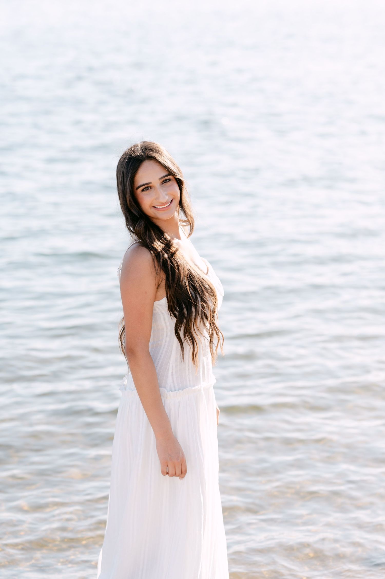 Young woman in white strapless maxi dress standing by sparkling ocean water, smiling gracefully at camera.