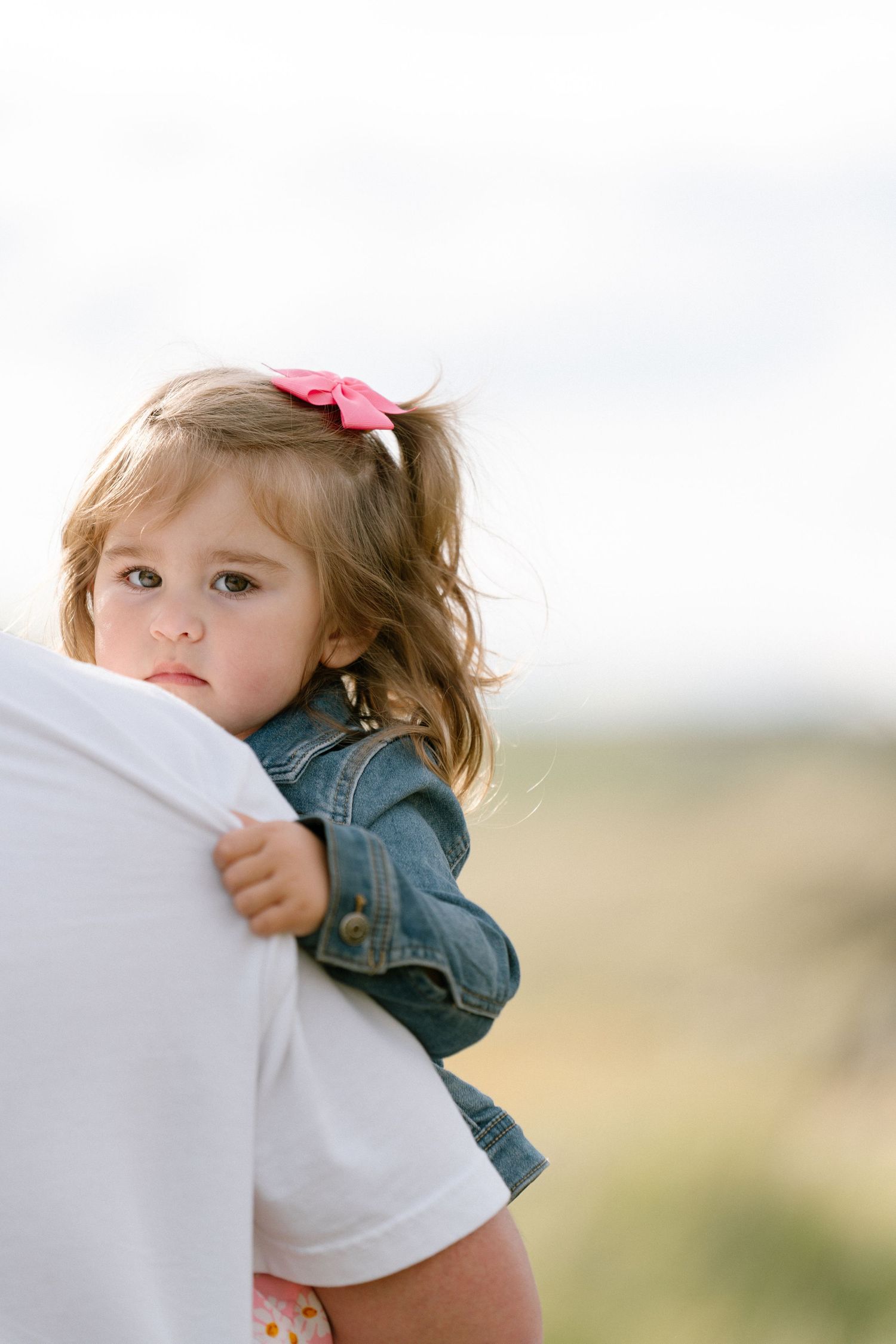 A toddler in a denim jacket with a pink bow being held against a blurred outdoor background.