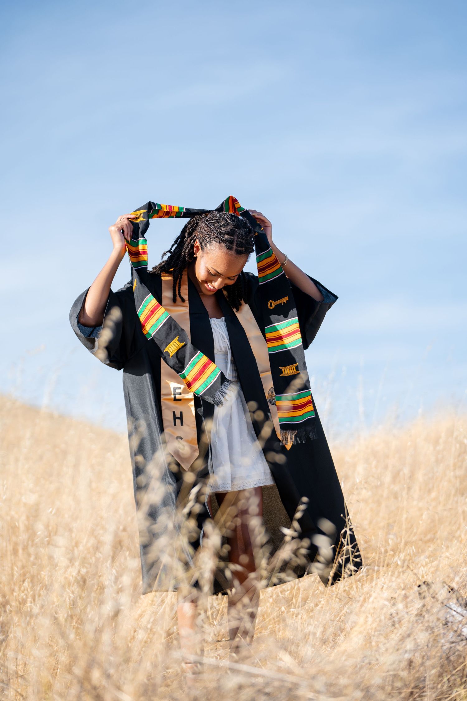 Person wearing graduation stole with colorful kente cloth pattern poses in tall grass field.