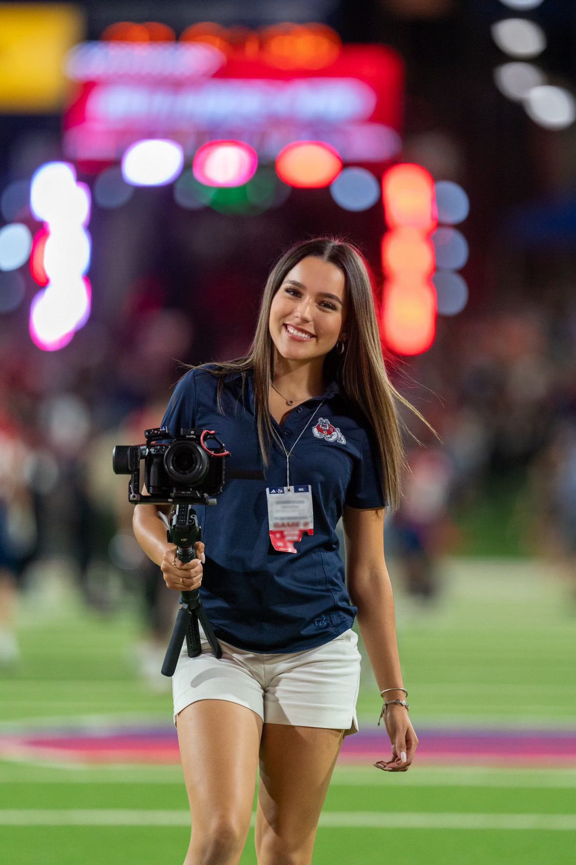 Camera operator in navy blue USA shirt and white shorts smiles while filming at a sports venue with colorful lights in background.