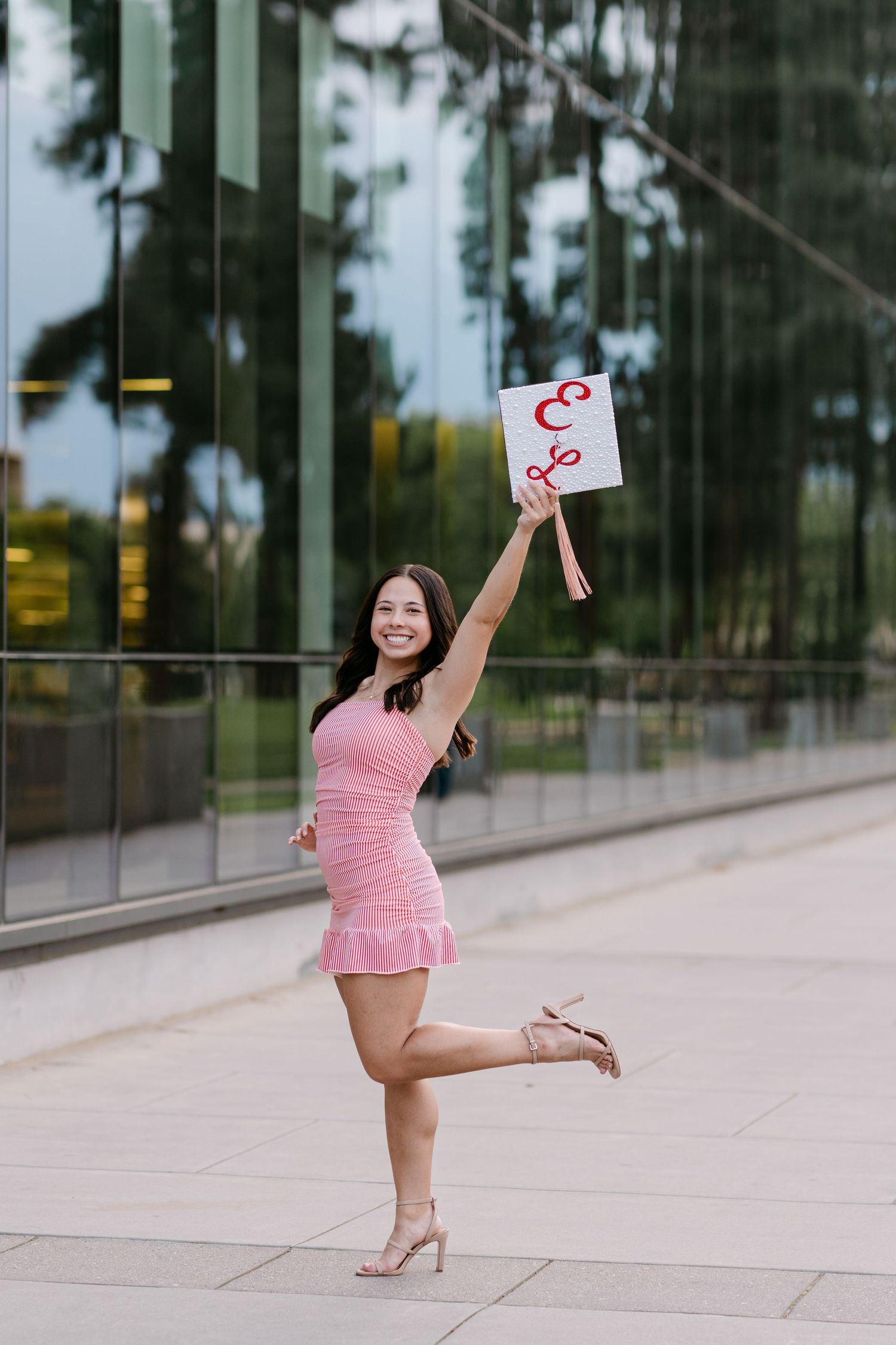 Young woman in pink dress celebrating outdoors, holding a sign up high while posing on one leg near glass building.