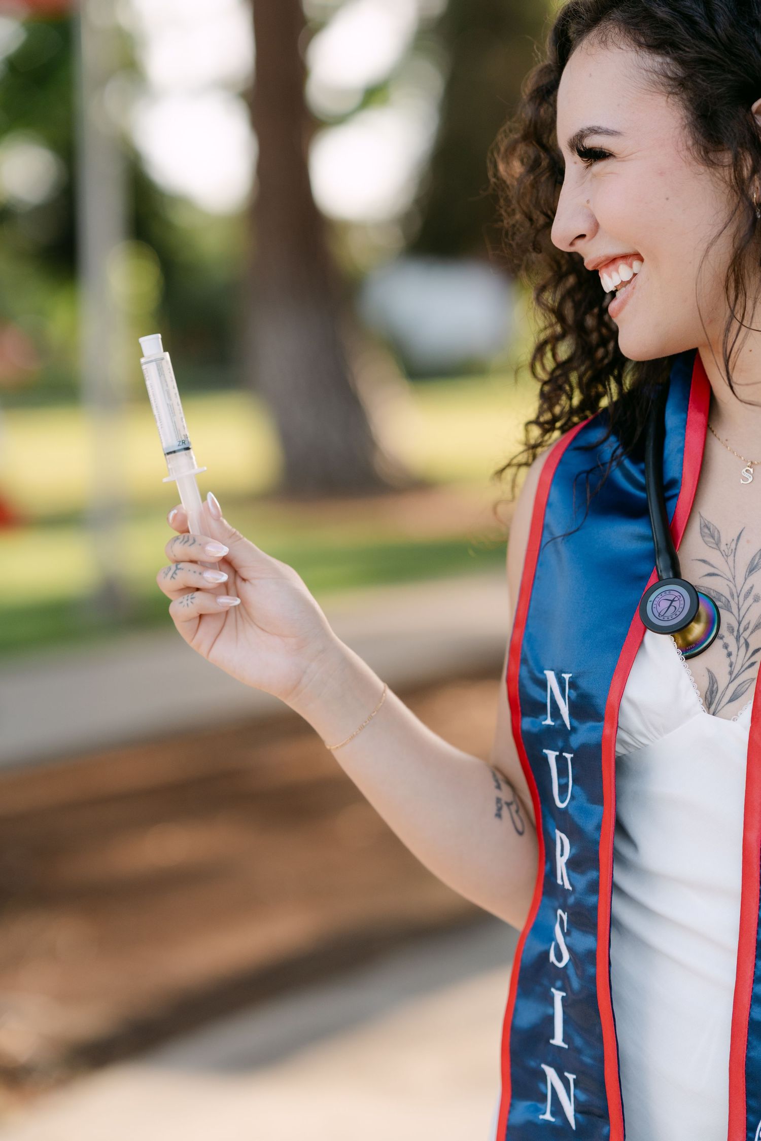 Happy nursing graduate holding a syringe outdoors wearing a blue NURSING graduation sash.