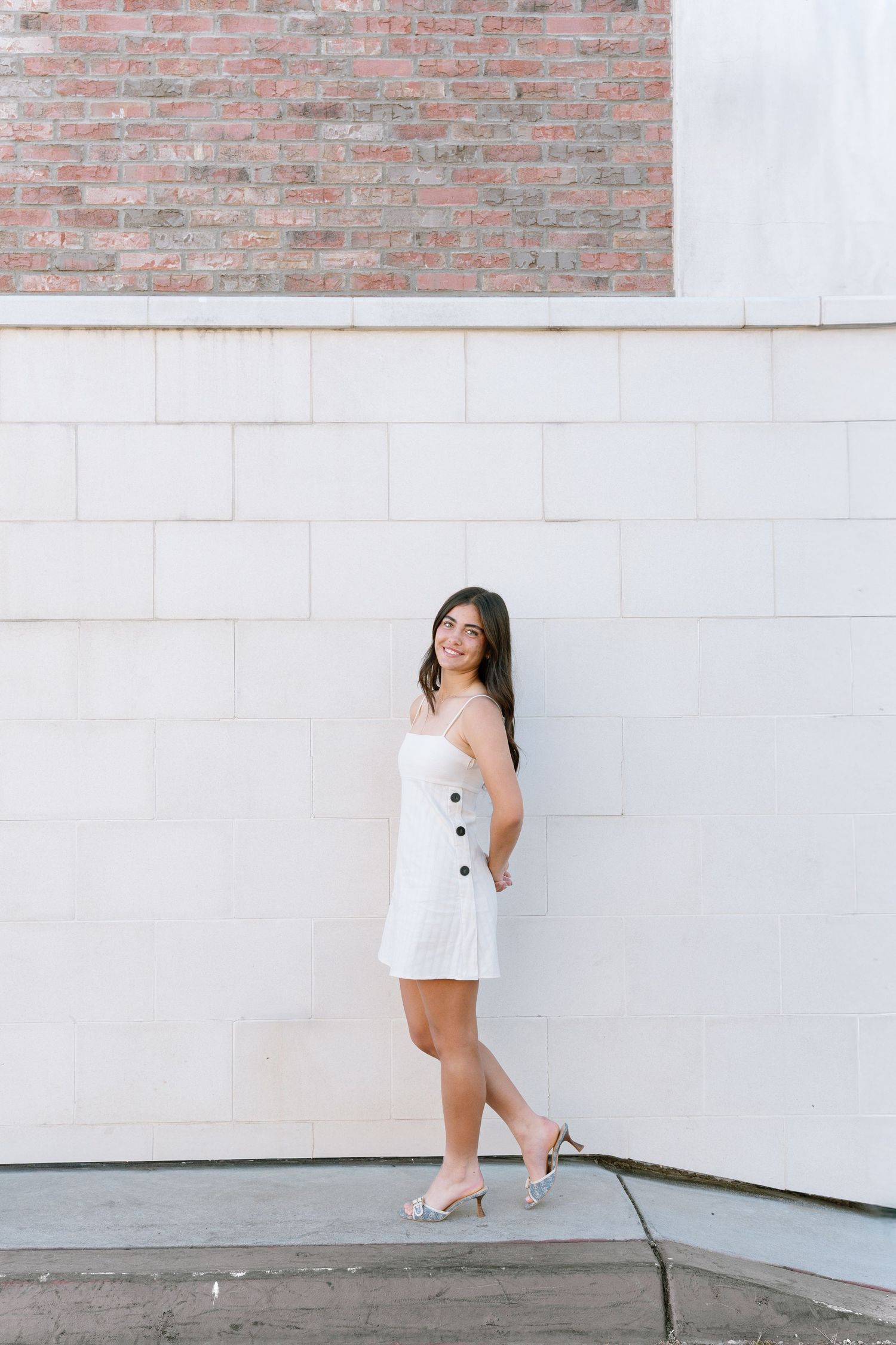 A person in a white outfit poses against a white wall with brick accents in an urban setting.